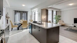 A modern kitchen with a central island, dining area with chairs, and a TV in the living room. - Sassafras Oasis | Sleeps 14 | All-Season Retreat Near Barrie (Minesing)