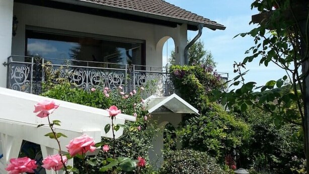 A two-story house with a balcony, surrounded by lush greenery and flowering plants. - 2 Bedroom Apartment in Bad Marienberg (Westerwald) (Bad Marienberg (Westerwald))