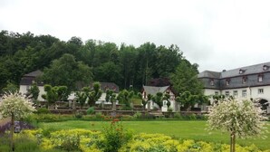 A large estate with multiple buildings, a well-maintained garden with flowering plants, and a forested hill in the background. - 2 Bedroom Apartment in Bad Marienberg (Westerwald) (Bad Marienberg (Westerwald))