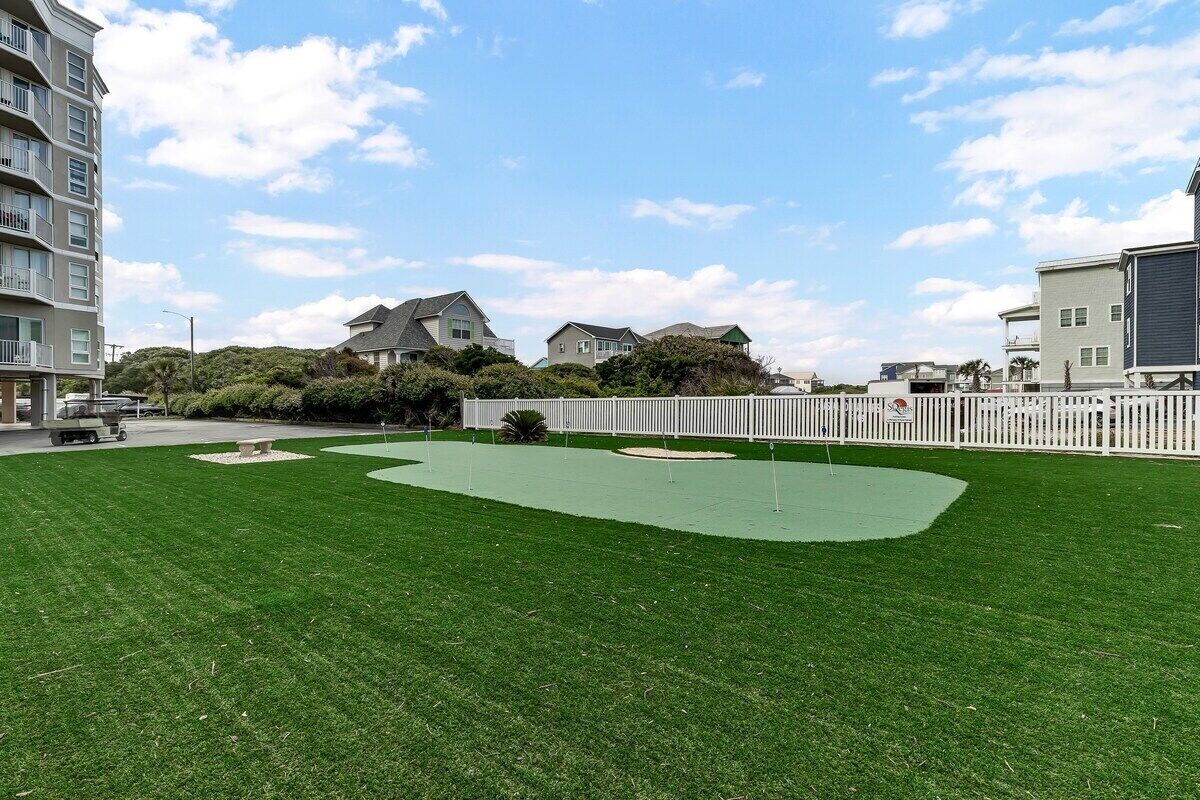 A well-maintained grassy area with a putting green, surrounded by a white fence and residential buildings in the background.
