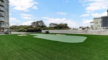 A well-maintained grassy area with a putting green, surrounded by a white fence and residential buildings in the background.