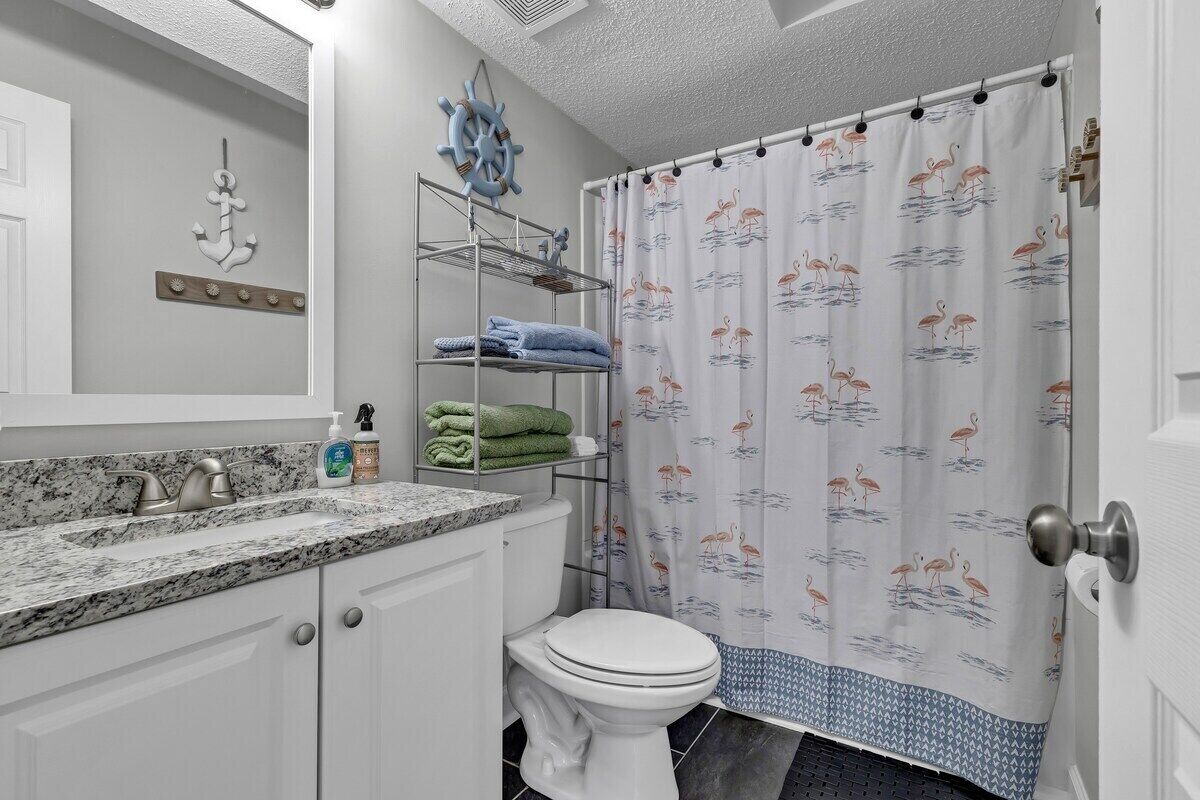 Une salle de bain avec des toilettes blanches, un lavabo doté d’un plan-vasque en marbre, un porte-serviettes avec des serviettes pliées et un rideau de douche orné d’un motif de flamants roses.