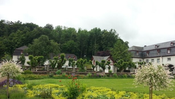 A large estate with multiple buildings, a well-maintained garden with various plants and flowers, and a forested hill in the background.