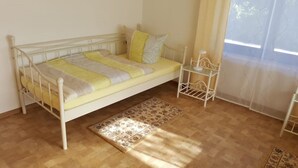 A single-bed room with a wooden floor, a patterned rug, and a window with sheer curtains.