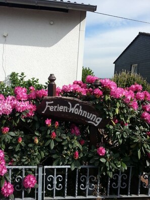 A wooden sign with the text "Ferienwohnung" in front of a white building, surrounded by pink flowers and a metal fence.