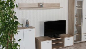 Ett modernt vardagsrum med en platt-TV på ett träställ, vita skåp, ett glasskåp och en krukväxt.