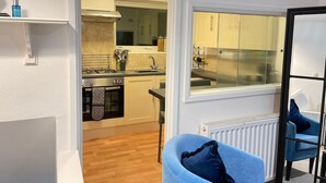Eine kleine Küche mit Gaskochfeld, Backofen und Spüle, ein Essbereich mit blauem Stuhl und ein Fenster mit Vorhängen.