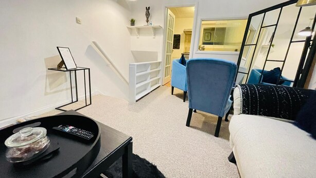 Ein kompakter Wohnbereich mit einer Couch, einem kleinen Tisch mit Fernbedienung, einem Sessel und einer Treppe zu einem angrenzenden Zimmer.