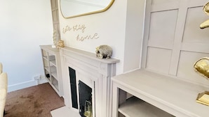 Une pièce avec une cheminée blanche, un miroir à cadre doré et une étagère ornée d’objets décoratifs.