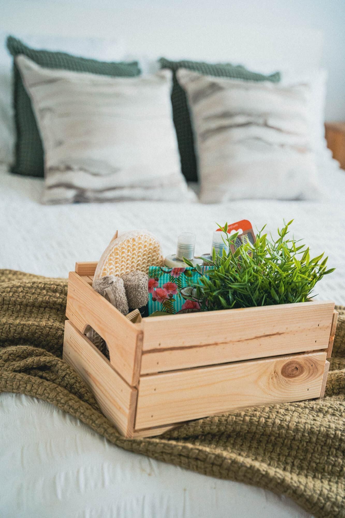 A wooden crate with a plant, a book, and a candle on a bed with white bedding and green pillows.