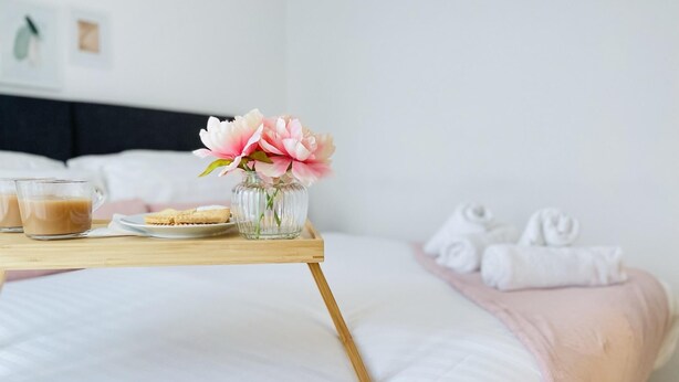 Ein ordentlich bezogenes Bett mit einer rosafarbenen Decke, ein Tablett mit zwei Gläsern Getränken und einem Teller Kekse, sowie eine Vase mit rosa Blumen.
