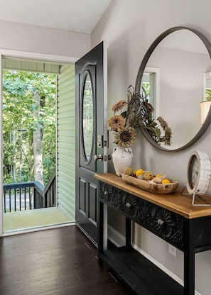 A hallway with a wooden bench, a vase with flowers, a clock, and a door leading to a balcony with a view of trees.