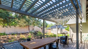 Eine Außenpatio mit Holztisch und -stühlen, einem darüberliegenden Pergola und Blick auf einen Garten mit Zaun und Bäumen.