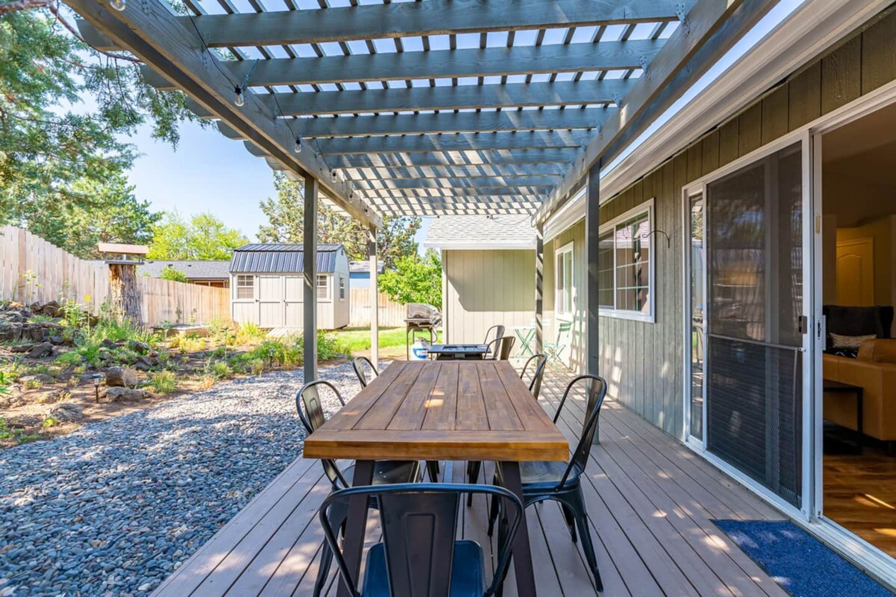 A covered patio with a wooden table and chairs, a pergola overhead, and a sliding glass door leading to an interior space.