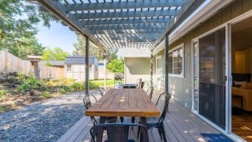 A covered patio with a wooden table and chairs, a pergola overhead, and a sliding glass door leading to an interior space.