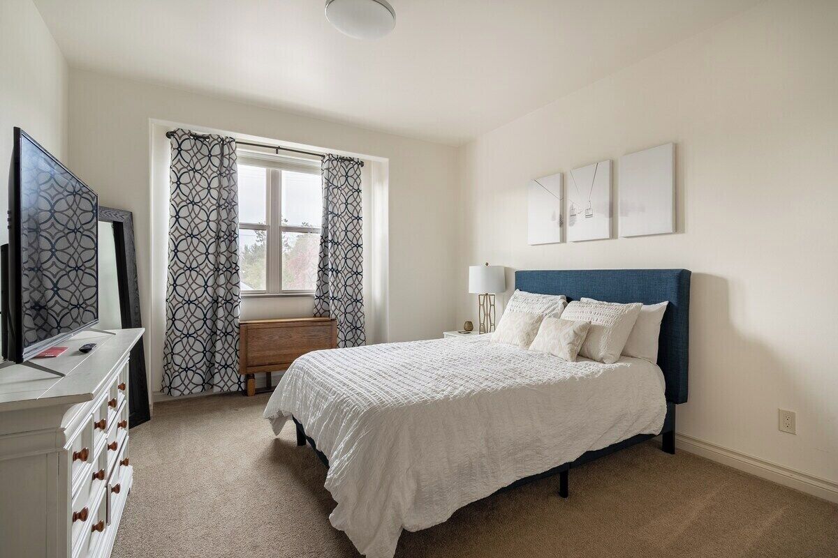 A bedroom with a bed, a dresser, a nightstand, a lamp, and a window with patterned curtains.