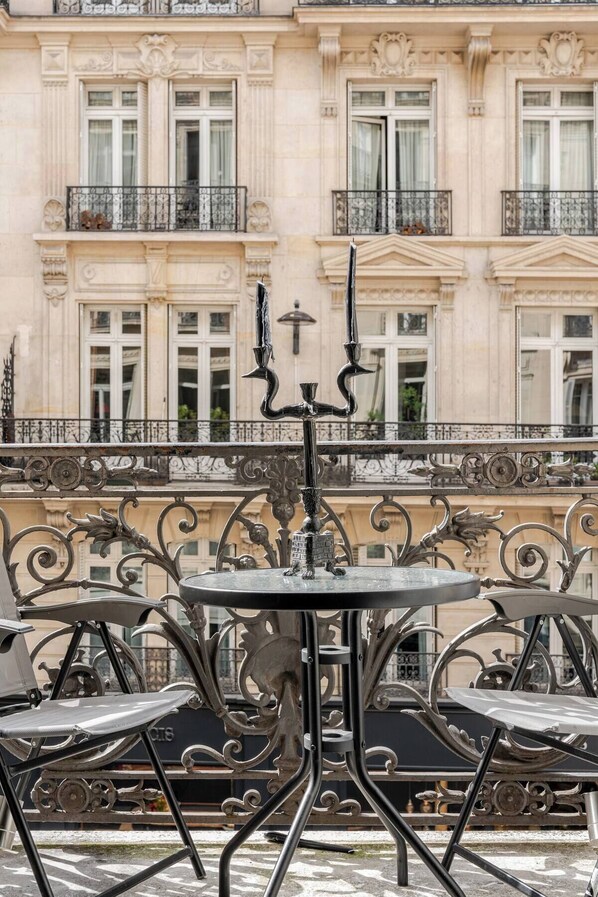 A wrought iron balcony railing with a table and chairs, a candelabra, and a building with classical architecture in the background.