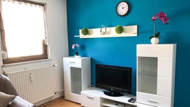 A living room with a blue wall, a white entertainment unit, a television, and a window with curtains. - 2 Bedroom Apartment in Bad Marienberg (Westerwald) (Bad Marienberg (Westerwald))