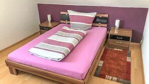 A single bed with a pink mattress and a striped blanket, flanked by wooden nightstands with lamps, in a room with wooden flooring and a purple accent wall. - 2 Bedroom Apartment in Bad Marienberg (Westerwald) (Bad Marienberg (Westerwald))