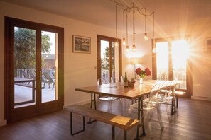 Een eetruimte met een houten tafel, transparante stoelen en een bank. Er zijn glazen deuren die leiden naar een terras met stoelen en een tafel.