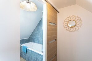 Een badkamer met een badkuip, een douche en een handdoek aan de badkuip. Er is een houten deur en een spiegel met een ronde vorm, bevestigd aan de muur.