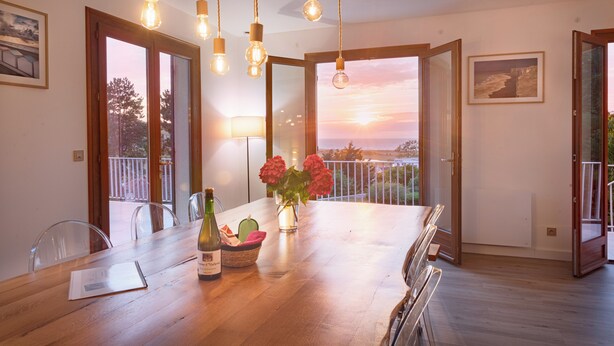 A dining area with a wooden table, chairs, hanging light fixtures, a vase with flowers, and a view of the sunset through open doors. - La Villa Maxine, sea view and spa (Criel-sur-Mer)