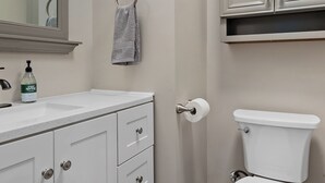 Ett badrum med ett vitt handfat, en spegel och en toalett. Det finns en handdukshängare med en handduk på, och en flaska med flytande tvål på handfatet. Väggarna är målade i en ljus beige färg, och det finns ett skåp ovanför toaletten.