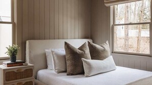 A bedroom with a wooden ceiling, a bed with white bedding, patterned pillows, a bedside table with a plant, and a window with a view.