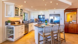 A modern kitchen with white cabinetry, a central island with bar stools, built-in appliances, and a wooden floor. - Polo Beach Club 106 2 Bedroom Condo (Kihei)