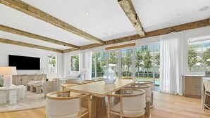 A spacious living room with a large wooden table, comfortable seating, and large windows offering a view of the outdoors.