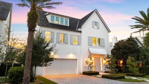 A two-story house with a white exterior, a garage, and a well-maintained garden with palm trees and shrubs.