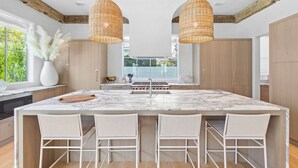 A modern kitchen with a large island, white wicker pendant lights, and a view of greenery outside.
