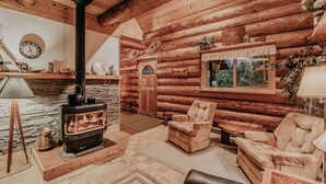 A log cabin interior with a stone fireplace, wooden furniture, and a large window overlooking greenery. - 11sl - Wi-fi - No Pets Log Cabin - Sleeps 7 2 Bedroom Cabin by RedAwning (Deming)