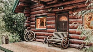 A rustic log cabin with a wooden porch, a bench, two large wooden wheels, and a door with a number '136' above it. - 11sl - Wi-fi - No Pets Log Cabin - Sleeps 7 2 Bedroom Cabin by RedAwning (Deming)