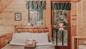 A cozy wooden cabin bedroom with a bed, a small table, a window with curtains, and a framed picture on the wall. - 11sl - Wi-fi - No Pets Log Cabin - Sleeps 7 2 Bedroom Cabin by RedAwning (Deming)