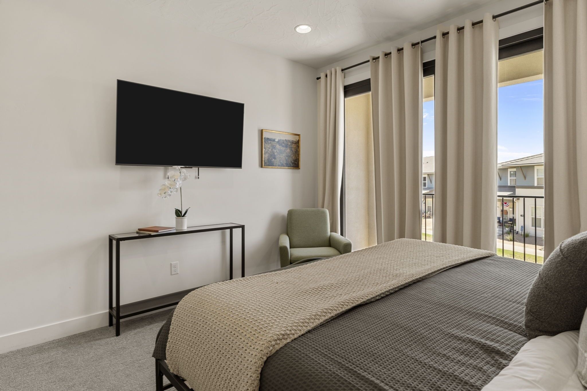 A hotel room with a bed, a TV mounted on the wall, a small table with a plant, a chair, and a window with curtains.