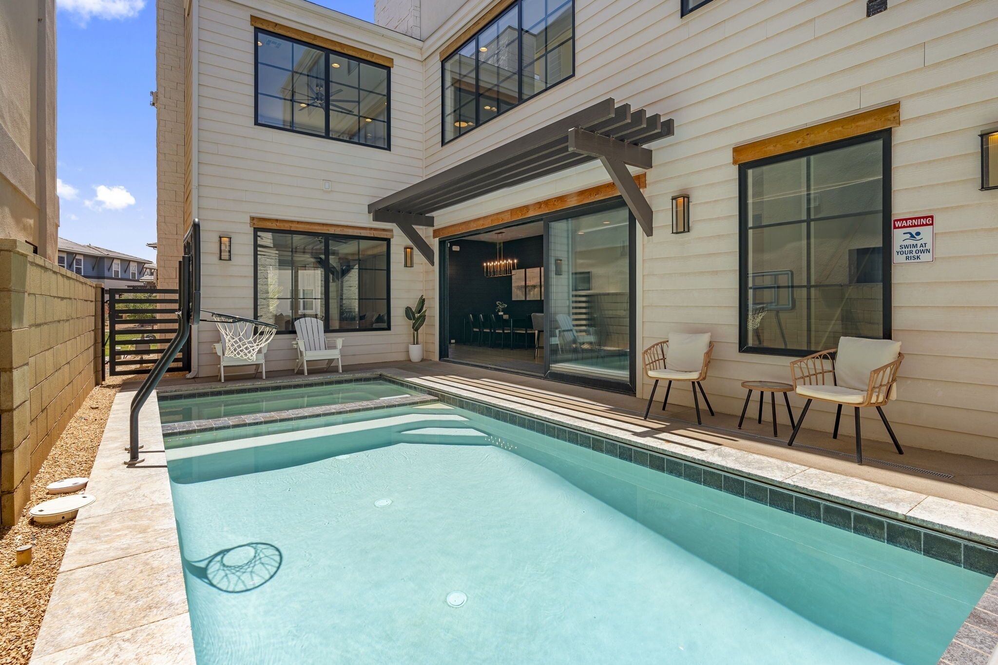 Piscina con sillas y una pérgola, un patio y un letrero de advertencia.