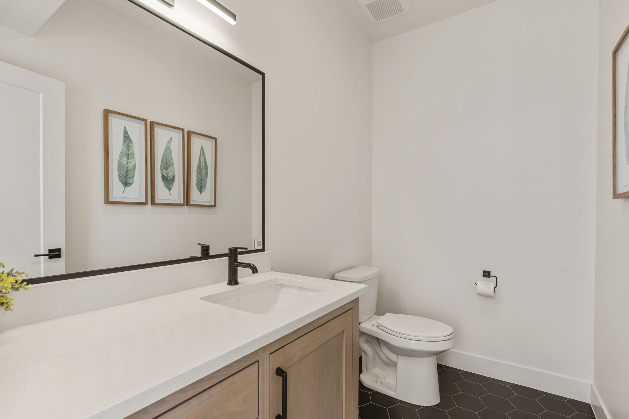 Un bany modern amb un lavabo blanc, un vàter i una obra d'art amb fulles emmarcades a la paret.