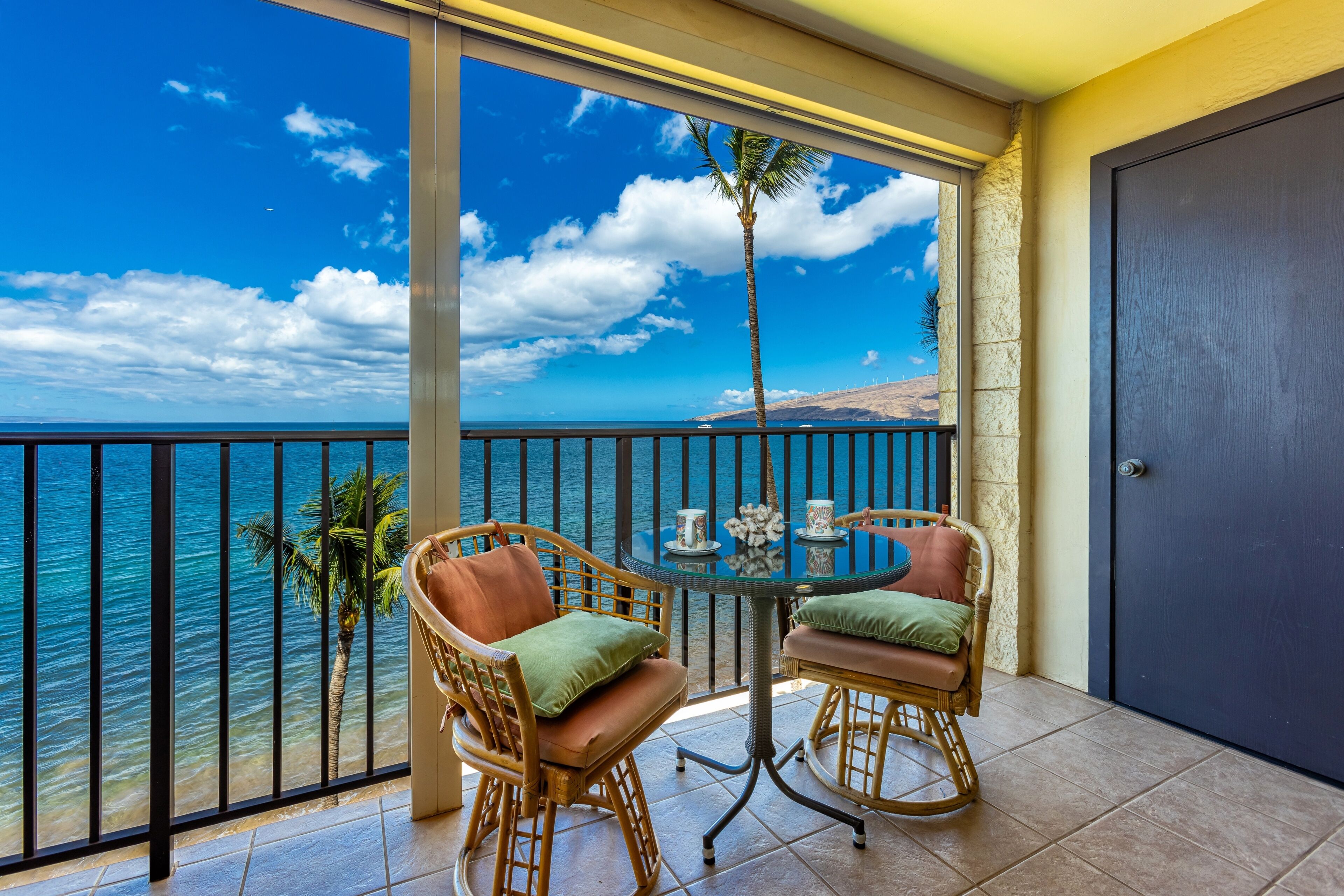 Un balcon avec vue sur l’océan, deux fauteuils en osier, une petite table ronde et une porte.