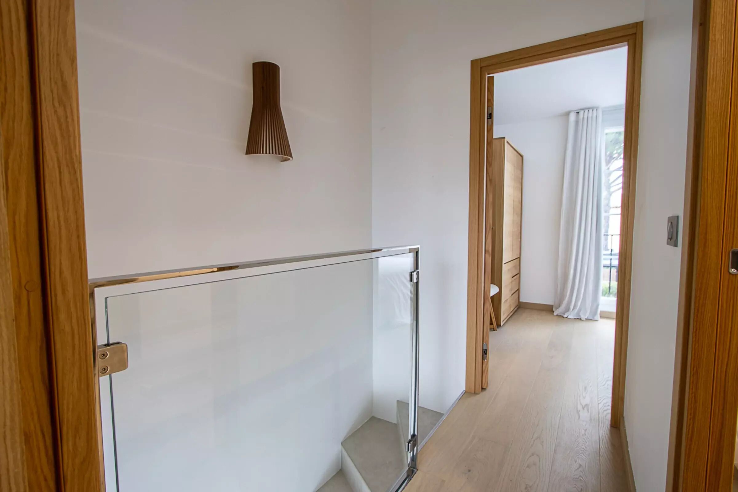 A hallway with a glass railing, wooden doors, and a view into a room with a white curtain and a wooden cabinet.