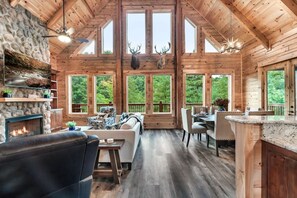 A cabin interior with a stone fireplace, wooden ceiling, large windows, and a dining area. - Brand New! Indoor heated pool, Mountain View’s! (Sevierville)