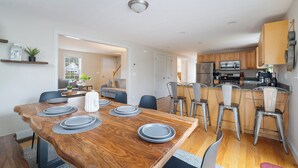 Eine moderne Küche mit Holzausstattung, einem integrierten Backofen und einem Edelstahlkühlschrank. Es gibt eine Essecke mit Holztisch und Stühlen, sowie eine Kücheninsel mit Barhocker.