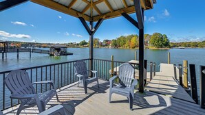 A covered deck with Adirondack chairs overlooking a lake, wooden dock, and distant buildings.