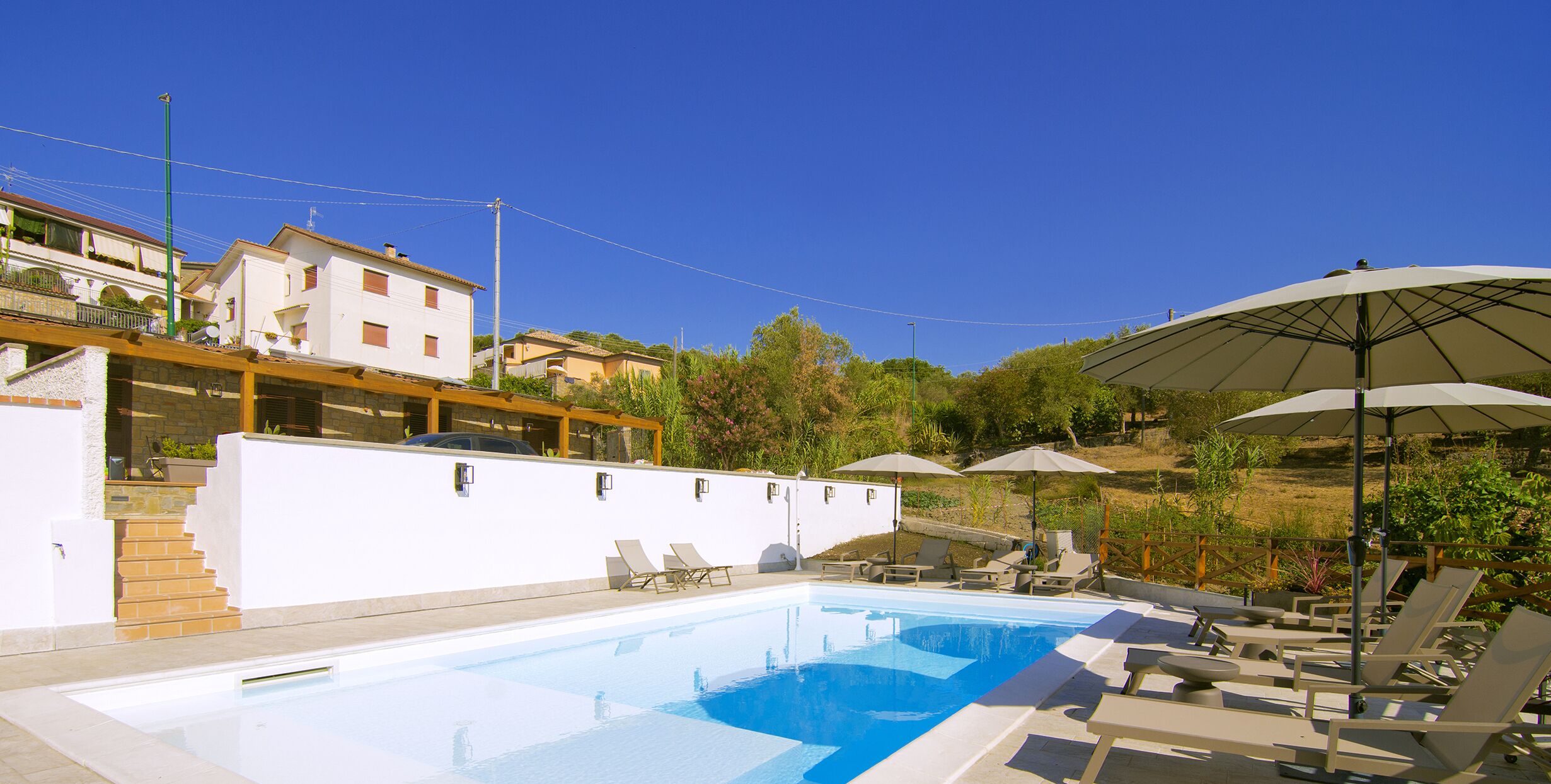 A swimming pool with lounge chairs and umbrellas, surrounded by buildings and greenery.
