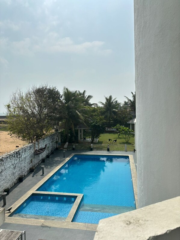 A rectangular swimming pool with a stone border, surrounded by a white wall and a stone wall. There are steps leading down to a grassy area with palm trees and a building labeled "CABANA".