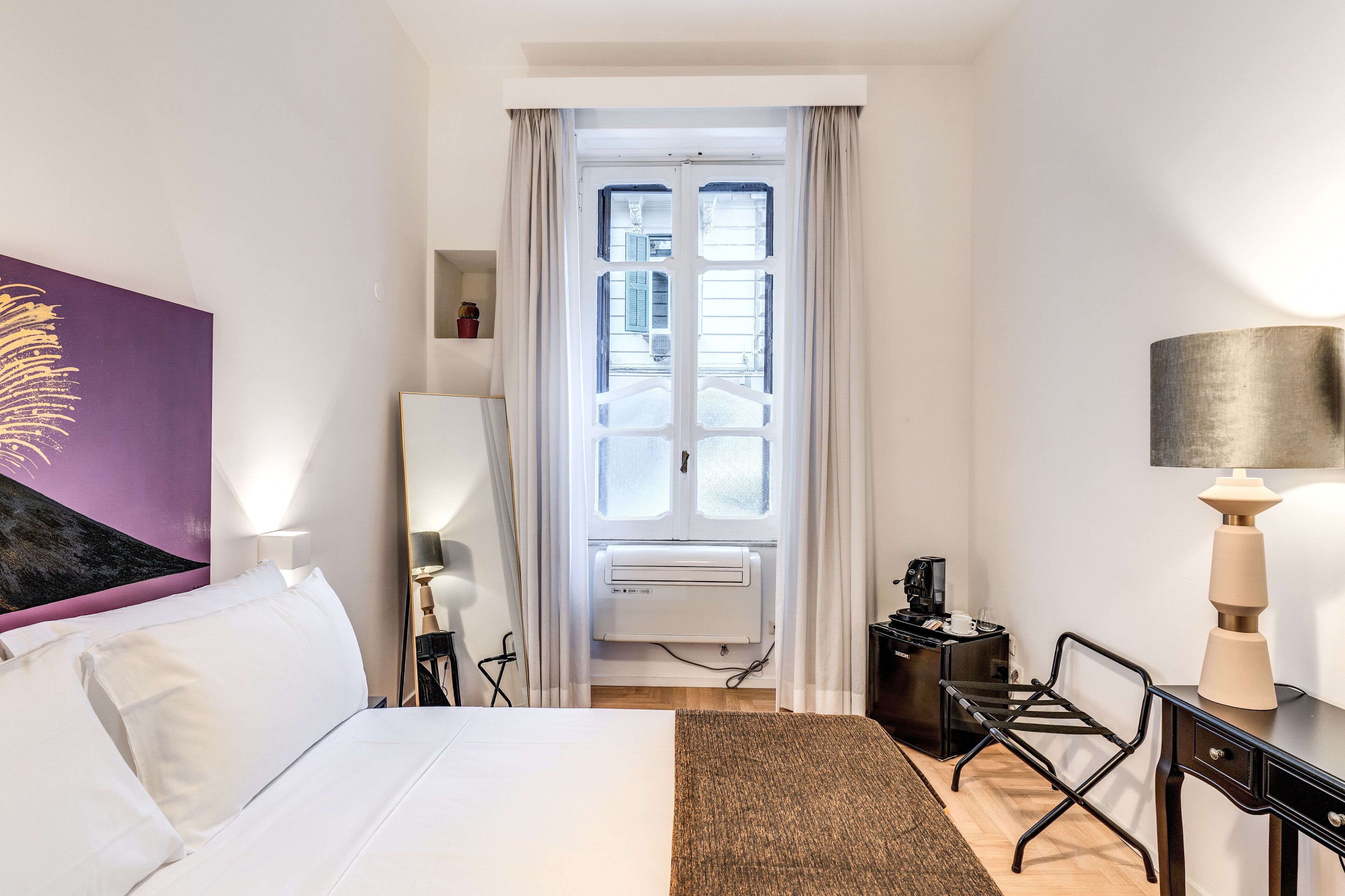 A hotel room with a bed, a desk, a chair, a lamp, a window with curtains, and a wall-mounted air conditioner.