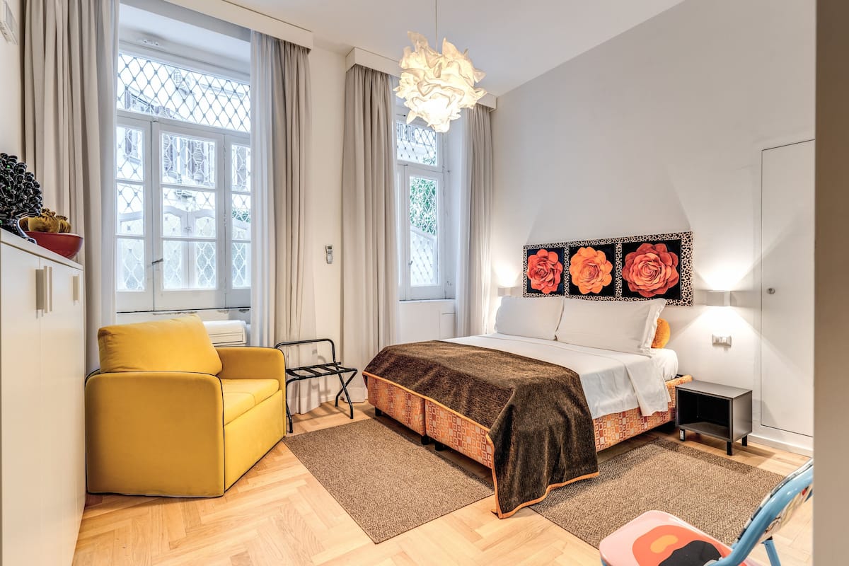 A bedroom with a bed, a yellow armchair, a small table, and a window with curtains.