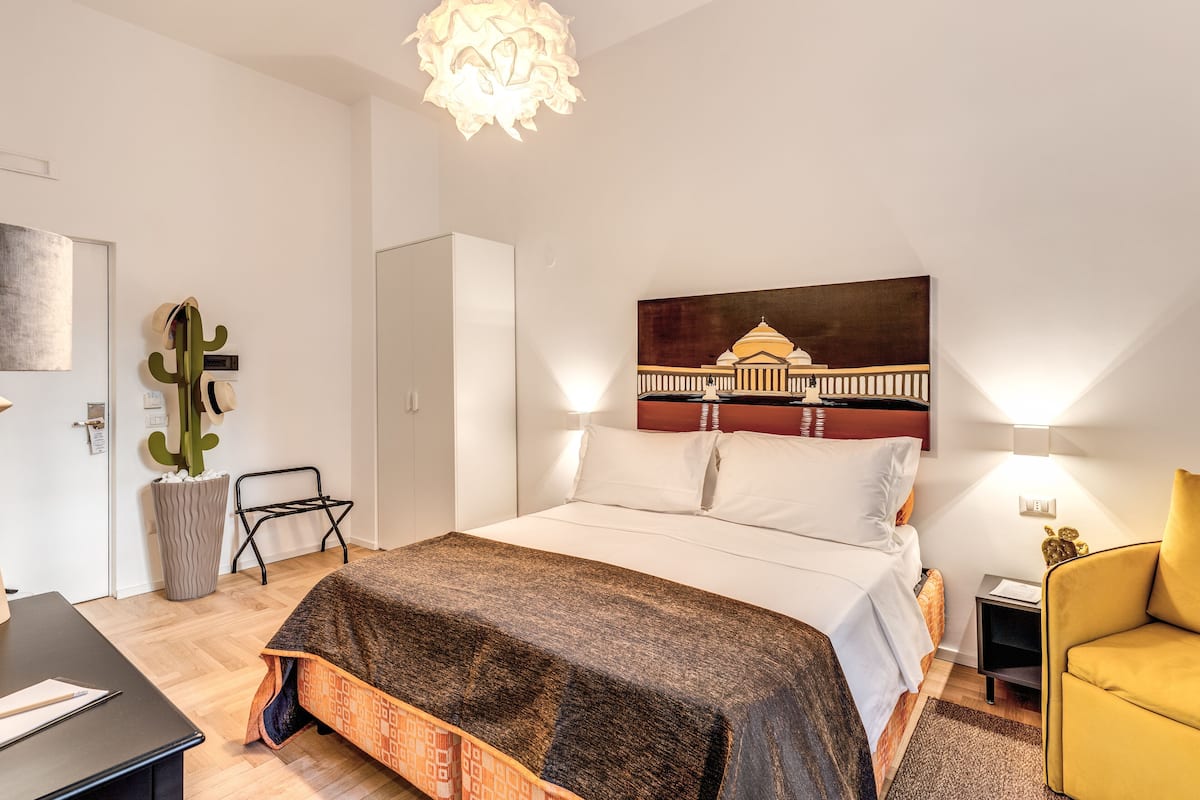 A modern bedroom with a large bed, a desk, a chair, a cactus, and a painting of a building.