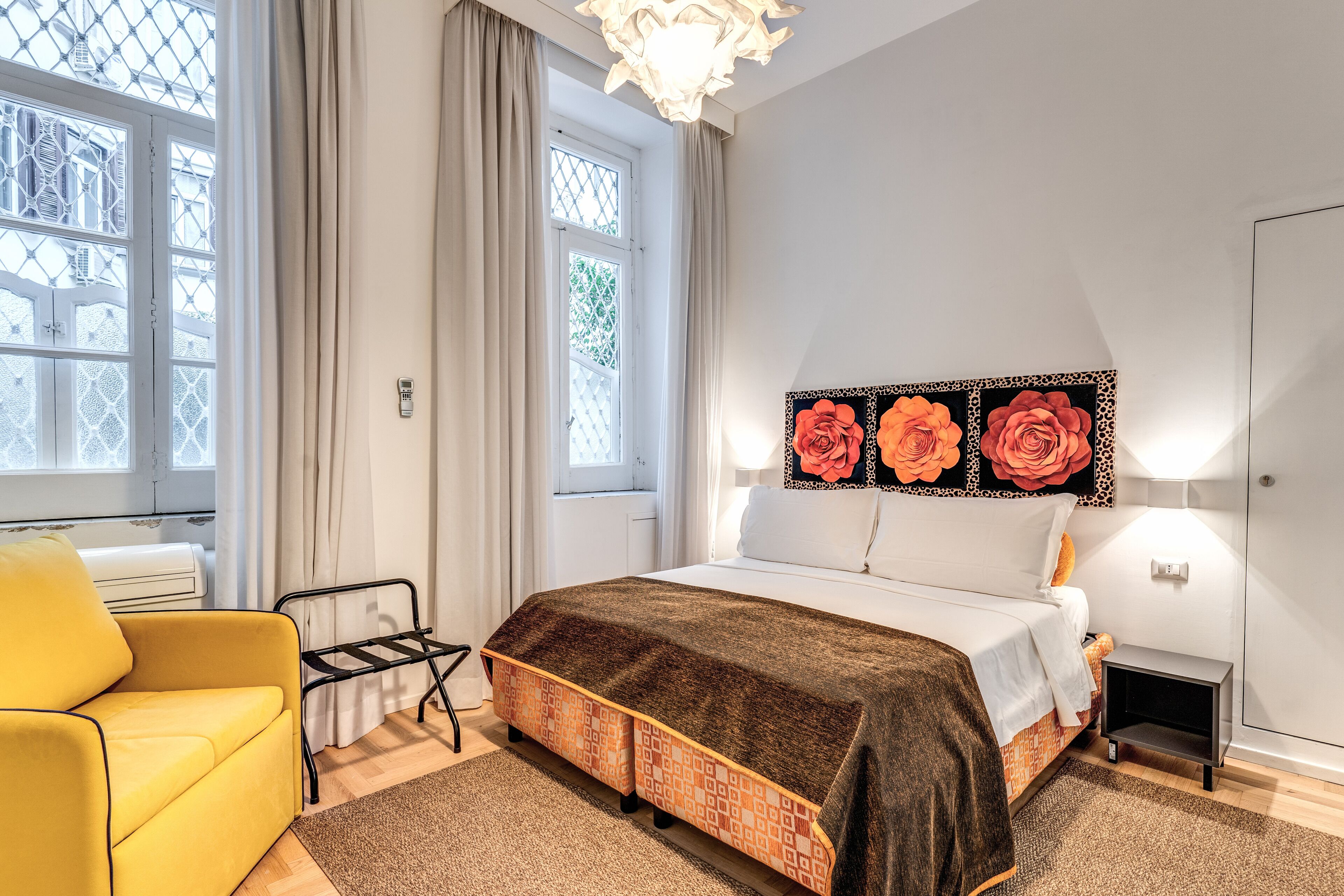 A bedroom with a bed, a yellow armchair, a radiator, a window with curtains, and a wall-mounted light fixture.