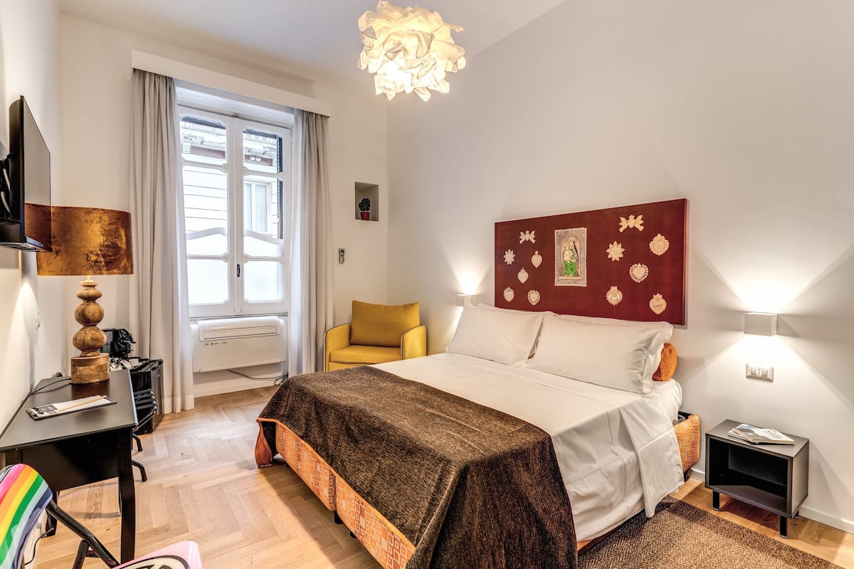 A hotel room with a bed, a desk, a chair, a lamp, a window, and a wall decoration.